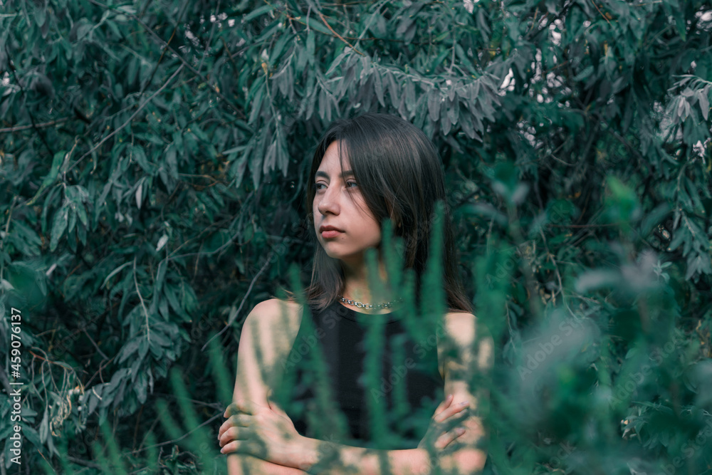 Naklejka premium A young brunette girl has her arms crossed over her chest and looks to the side against a background of dark green leaves. The girl is alone in the forest. High quality photo