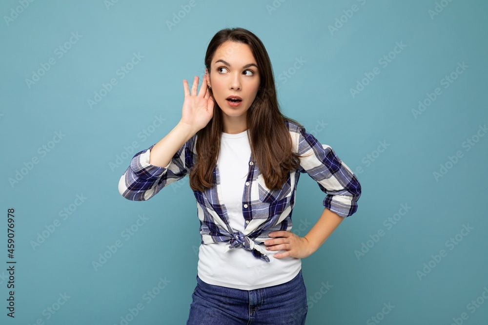 Photo of young concentrated attractive pretty brunet woman with sincere emotions wearing stylish check shirt standing isolated on blue background with empty space and listening to something by putting Photo of young concentrated attractive pretty brunet woman with sincere emotions wearing stylish check shirt standing isolated on blue background with empty space and listening to something by putting