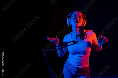 Caucasian woman in glasses and headphones sings into a microphone in neon light on a black background. An emotional girl is recording a song in a recording studio