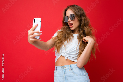 Sexy amazing beautiful young woman holding mobile phone taking selfie photo using smartphone camera wearing everyday stylish outfit and sunglasses isolated over colorful wall background looking at