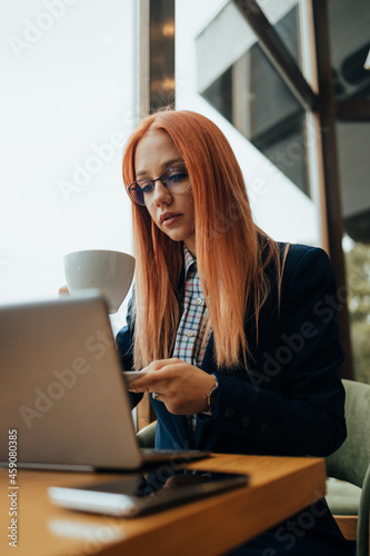 Young attractive redhead business woman or college student in a cafe bar or restaurant. She is sitting next to window, drinking coffee and working something on laptop computer.