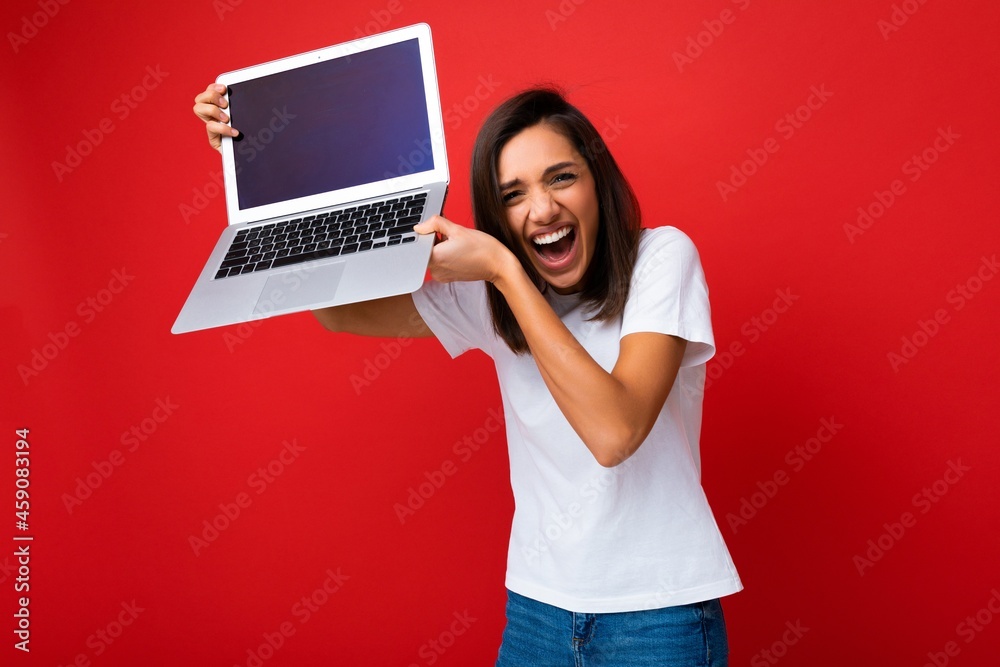 Photo of pretty Beautiful happy overjoyed young woman with short dark brunet haircut holding computer laptop up to head looking at camera wearing white t-shirt and jeans isolated over red wall Photo of pretty Beautiful happy overjoyed young woman with short dark brunet haircut holding computer laptop up to head looking at camera wearing white t-shirt and jeans isolated over red wall