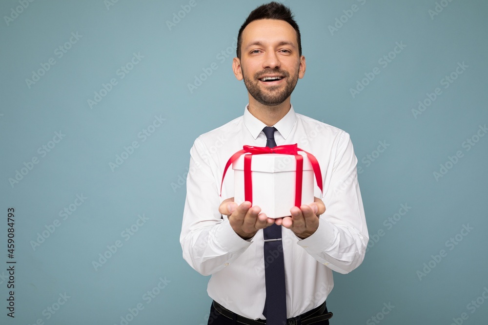 Photo shot of handsome positive brunette unshaven young man isolated over blue background wall wearing white shirt holding white gift box with red ribbon and looking at camera