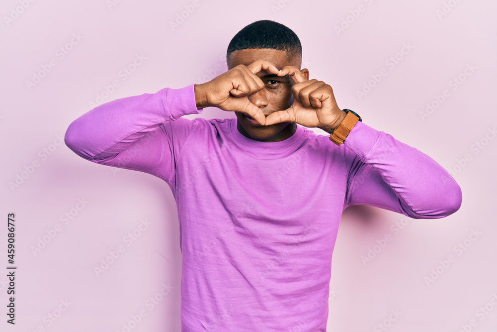 Young black man wearing casual pink sweater doing heart shape with hand and fingers smiling looking through sign Young black man wearing casual pink sweater doing heart shape with hand and fingers smiling looking through sign
