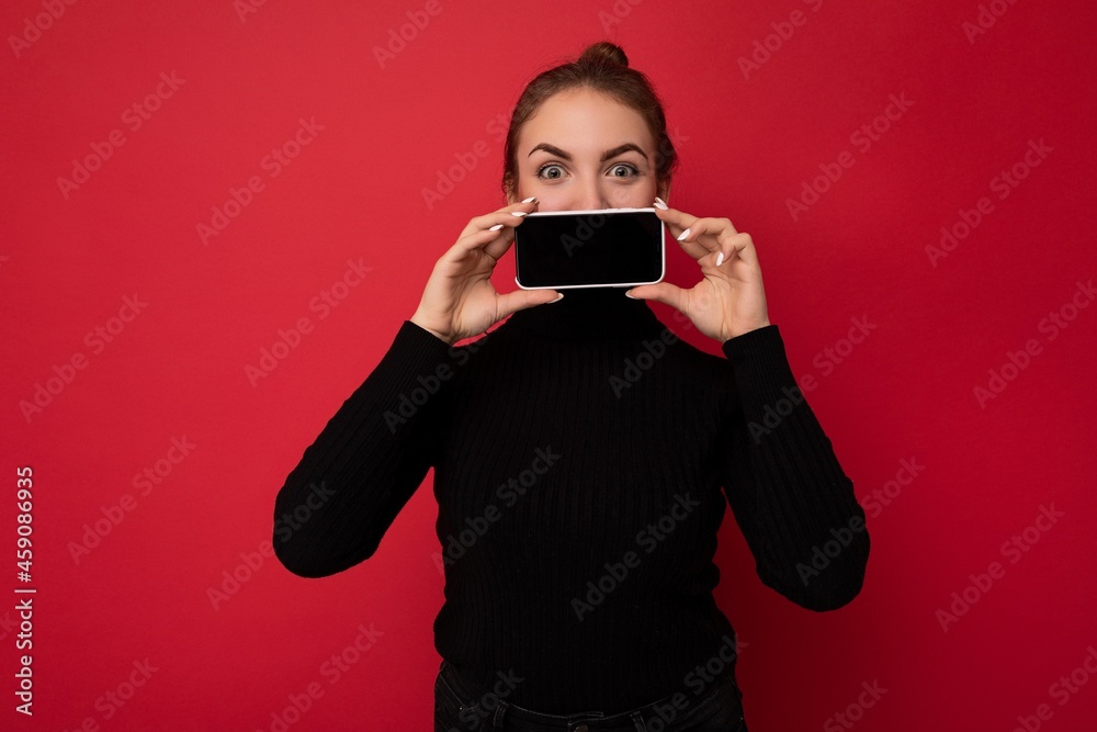 Photo of attractive positive young brunette woman wearing black sweater standing isolated over red background showing mobile phone with empty screen for mockup looking at camera Photo of attractive positive young brunette woman wearing black sweater standing isolated over red background showing mobile phone with empty screen for mockup looking at camera