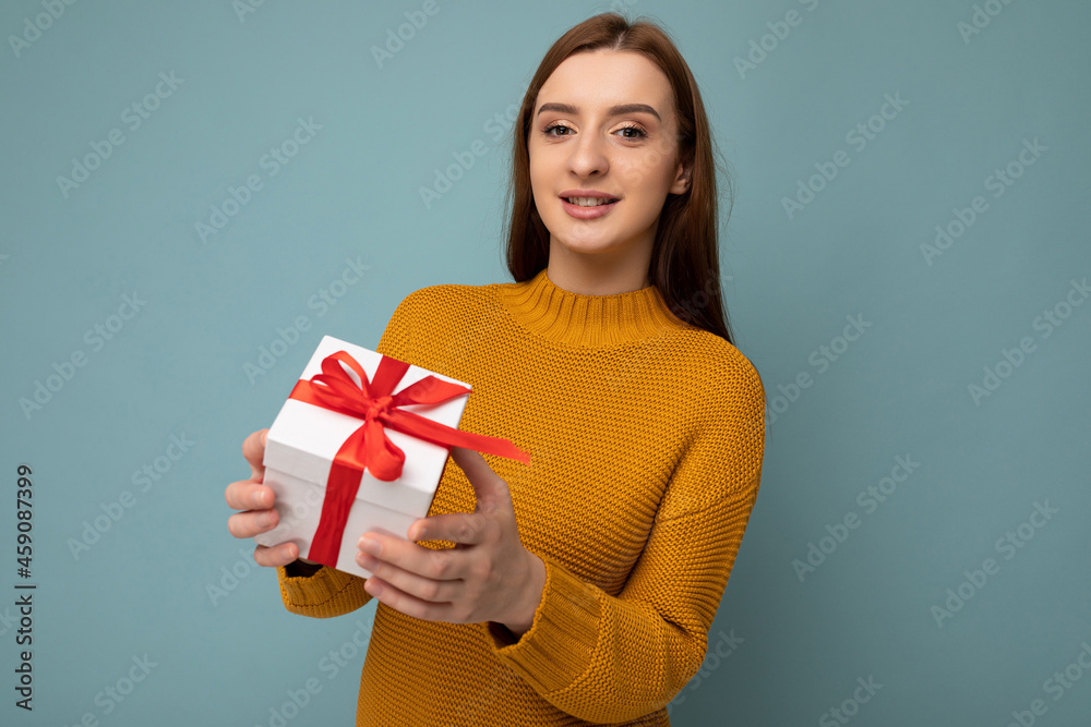 Beautiful happy young brunette woman isolated over colourful background wall wearing stylish casual clothes holding gift box and looking at camera