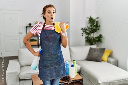 Young brunette woman wearing apron holding cleaning products at home surprised pointing with hand finger to the side, open mouth amazed expression.