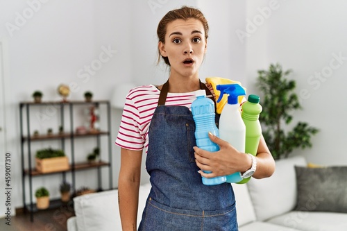 Young brunette woman wearing apron holding cleaning products scared and amazed with open mouth for surprise, disbelief face