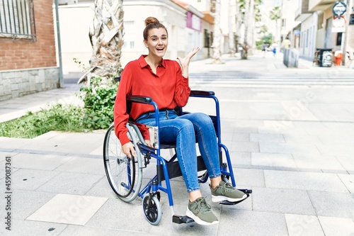 Young brunette woman sitting on wheelchair outdoors celebrating victory with happy smile and winner expression with raised hands