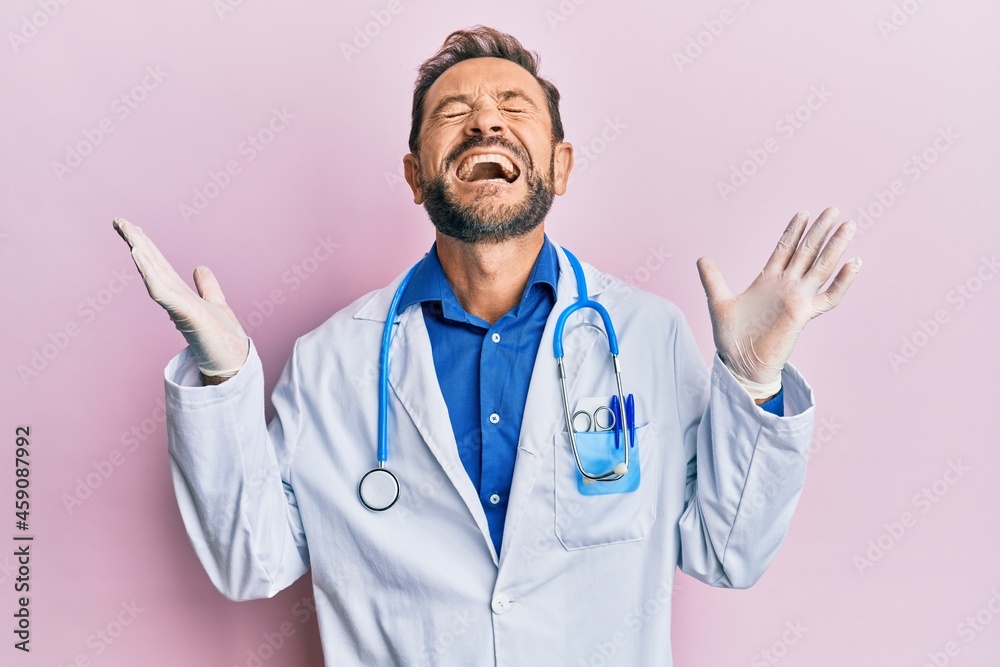 Middle age man wearing doctor uniform and stethoscope celebrating mad and crazy for success with arms raised and closed eyes screaming excited. winner concept Middle age man wearing doctor uniform and stethoscope celebrating mad and crazy for success with arms raised and closed eyes screaming excited. winner concept