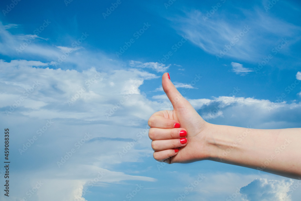 A woman making shows a like sign by holding her thumb up. Female hand with red gel polish manicure against the background of a blue sky with white clouds.. Copy space A woman making shows a like sign by holding her thumb up. Female hand with red gel polish manicure against the background of a blue sky with white clouds.. Copy space