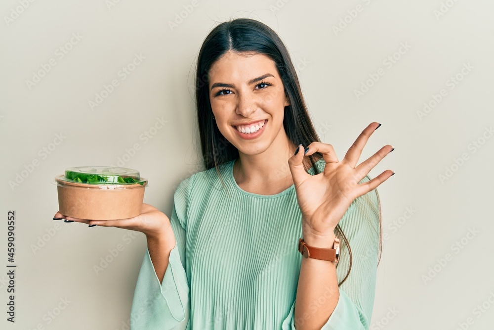 Young hispanic woman eating take away salad doing ok sign with fingers, smiling friendly gesturing excellent symbol Young hispanic woman eating take away salad doing ok sign with fingers, smiling friendly gesturing excellent symbol