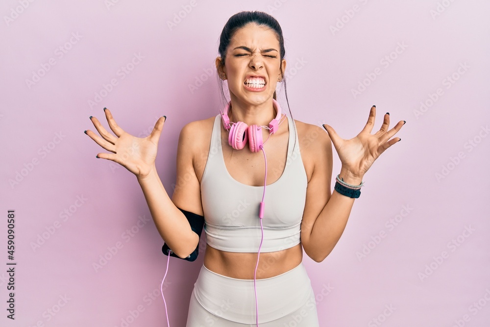 Young hispanic woman wearing gym clothes and using headphones crazy and mad shouting and yelling with aggressive expression and arms raised. frustration concept.