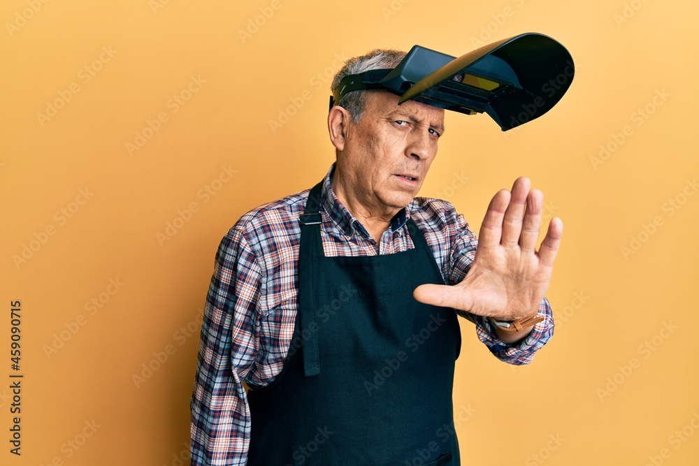 Handsome senior man with grey hair wearing welding protection mask doing stop sing with palm of the hand. warning expression with negative and serious gesture on the face. Handsome senior man with grey hair wearing welding protection mask doing stop sing with palm of the hand. warning expression with negative and serious gesture on the face.
