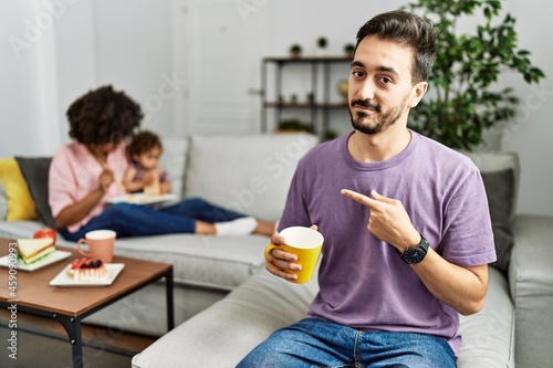 Hispanic father of interracial family drinking a cup coffee pointing with hand finger to the side showing advertisement  serious and calm face