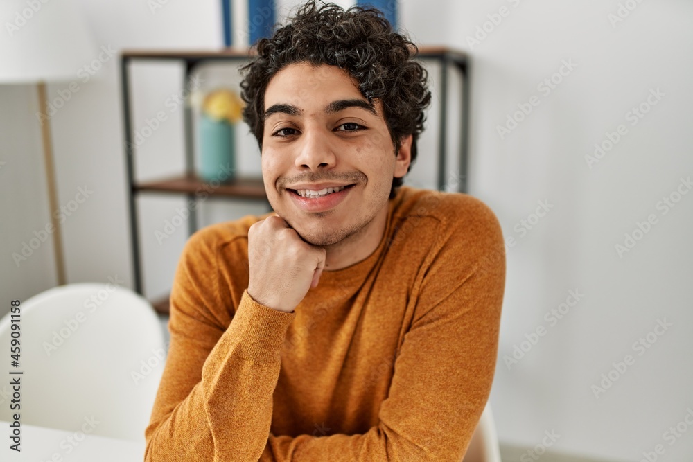 Young hispanic man smiling happy sitting on the table at home. Young hispanic man smiling happy sitting on the table at home.