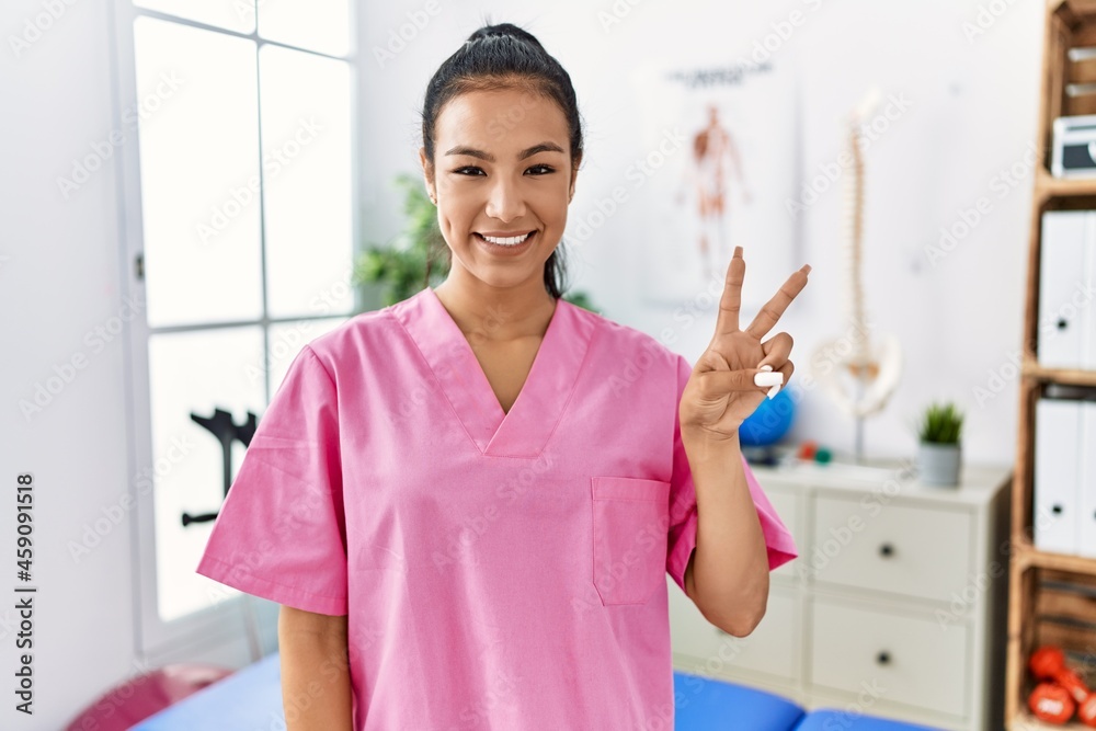Young hispanic woman working at pain recovery clinic showing and pointing up with fingers number two while smiling confident and happy. Young hispanic woman working at pain recovery clinic showing and pointing up with fingers number two while smiling confident and happy.