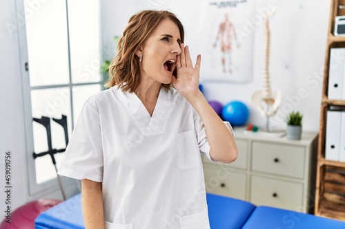 Middle age physiotherapist woman working at pain recovery clinic shouting and screaming loud to side with hand on mouth. communication concept.
