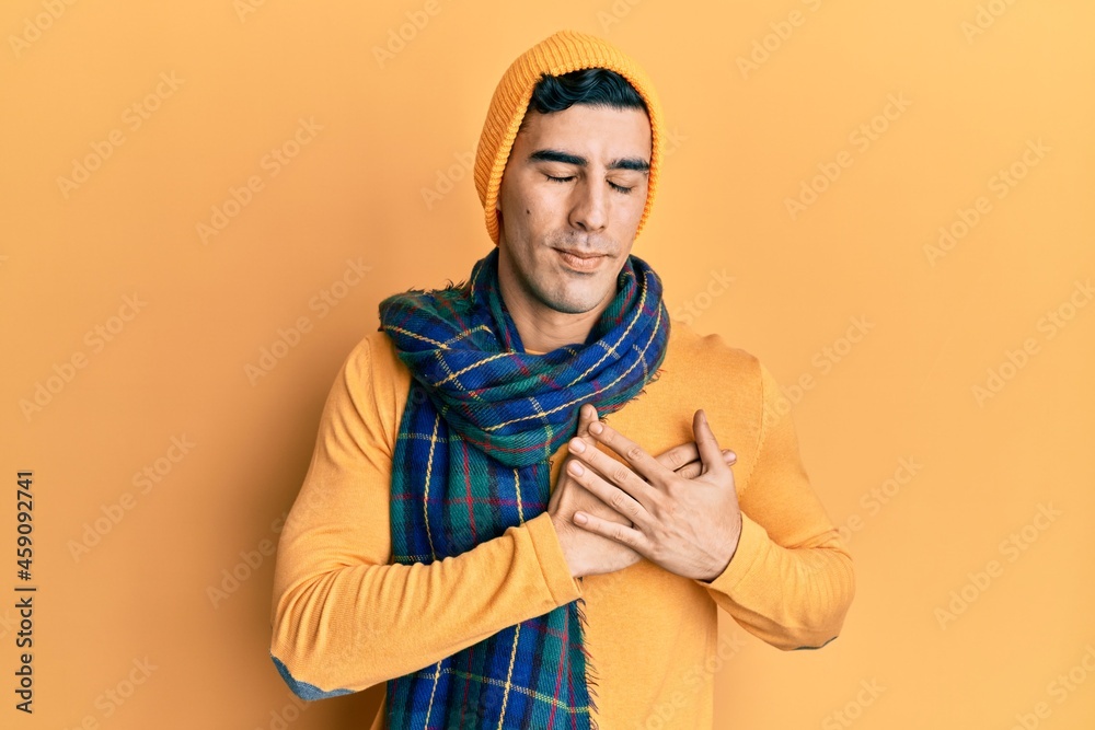 Handsome hispanic man wearing wool hat and winter scarf smiling with hands on chest with closed eyes and grateful gesture on face. health concept. Handsome hispanic man wearing wool hat and winter scarf smiling with hands on chest with closed eyes and grateful gesture on face. health concept.