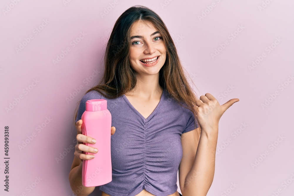 Young caucasian woman holding shampoo bottle pointing thumb up to the side smiling happy with open mouth Young caucasian woman holding shampoo bottle pointing thumb up to the side smiling happy with open mouth