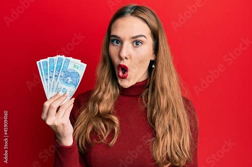 Young blonde woman holding 50 polish zloty banknotes scared and amazed with open mouth for surprise, disbelief face