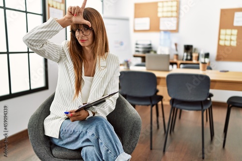 Hispanic business woman working at the office making fun of people with fingers on forehead doing loser gesture mocking and insulting. © Krakenimages.com