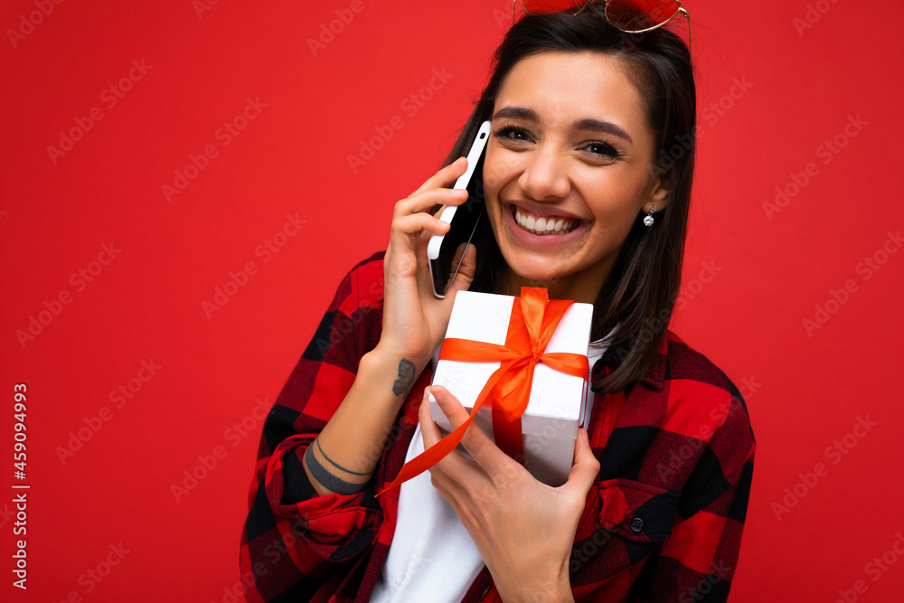 Shot of beautiful happy emotional young brunette woman isolated over red background wall wearing white casual t-shirt and red and black shirt holding white gift box with red ribbon and talking on Shot of beautiful happy emotional young brunette woman isolated over red background wall wearing white casual t-shirt and red and black shirt holding white gift box with red ribbon and talking on