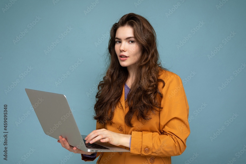Close-up portrait of Beautiful brunet curly young woman holding netbook computer looking at camera wearing yellow jacket typing on keyboard isolated on blue background Close-up portrait of Beautiful brunet curly young woman holding netbook computer looking at camera wearing yellow jacket typing on keyboard isolated on blue background