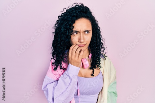 Young hispanic woman with curly hair wearing gym clothes and using headphones looking stressed and nervous with hands on mouth biting nails. anxiety problem.
