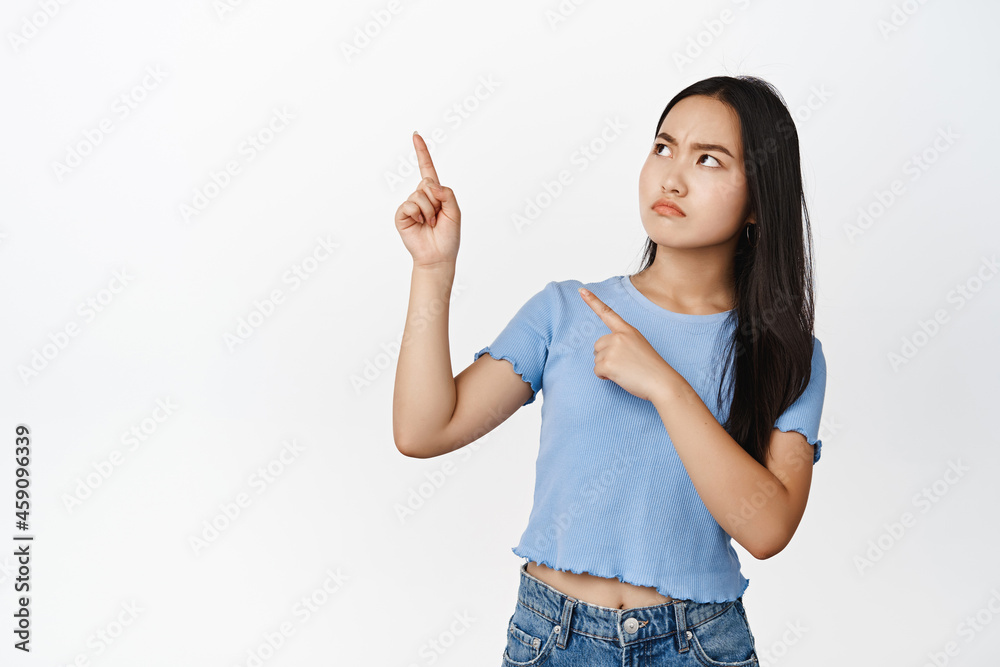 Displeased vietnamese girl pointing and looking at upper left corner, showing something concerning or bad, white background Displeased vietnamese girl pointing and looking at upper left corner, showing something concerning or bad, white background
