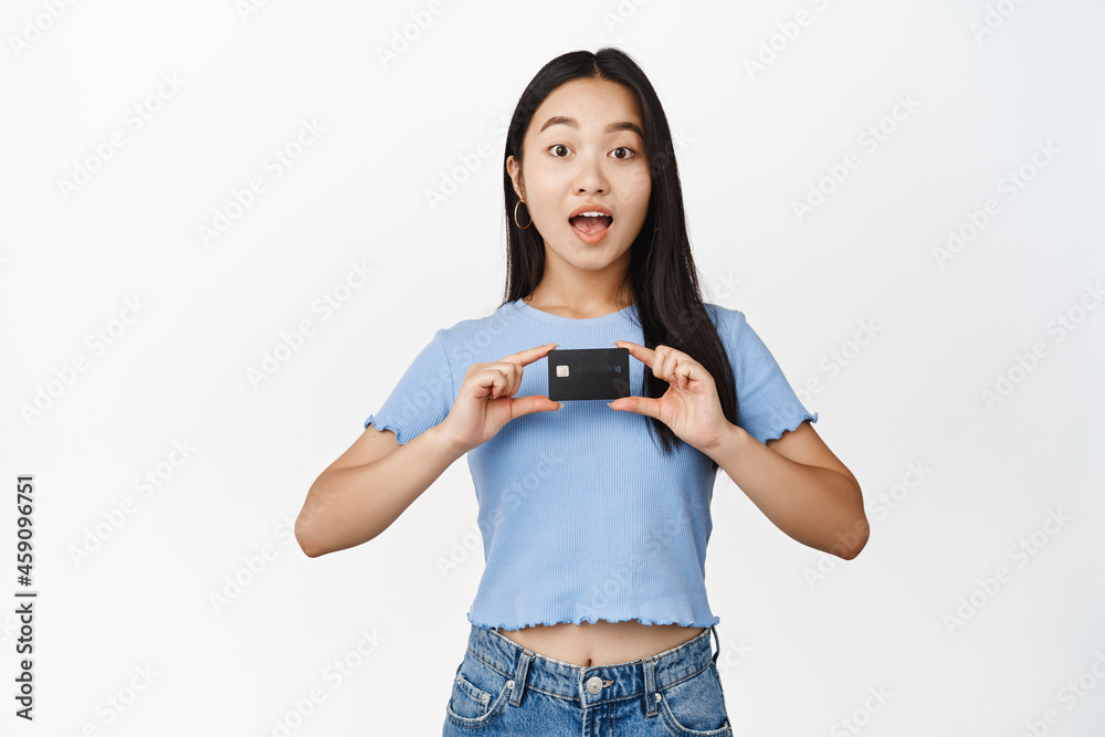 Image of young asian woman showing credit card, looking amazed at camera, standing in t-shirt over white background Image of young asian woman showing credit card, looking amazed at camera, standing in t-shirt over white background