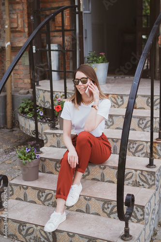 Stylish woman sitting on the steps of a building on the street and talking on the phone