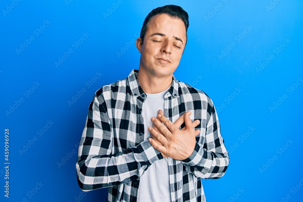 Handsome young man wearing casual shirt smiling with hands on chest, eyes closed with grateful gesture on face. health concept. Handsome young man wearing casual shirt smiling with hands on chest, eyes closed with grateful gesture on face. health concept.