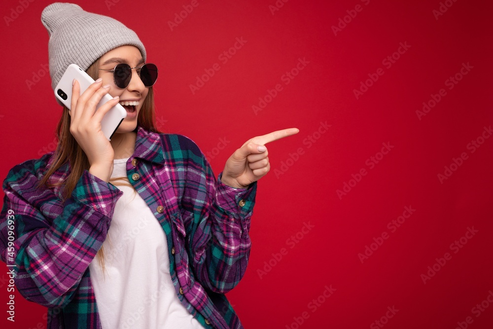 photo of beautiful happy positive young blonde woman wearing hipster purple shirt and casual white t-shirt grey hat and sunglasses isolated over red background holding in hand and talking on mobile photo of beautiful happy positive young blonde woman wearing hipster purple shirt and casual white t-shirt grey hat and sunglasses isolated over red background holding in hand and talking on mobile