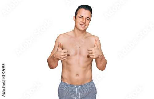 Handsome young man wearing swimwear shirtless success sign doing positive gesture with hand, thumbs up smiling and happy. cheerful expression and winner gesture.