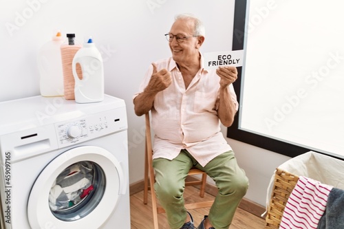 Senior man doing laundry holding eco friendly paper pointing thumb up to the side smiling happy with open mouth