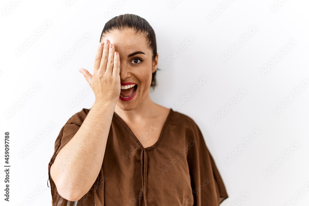 Young brunette woman standing over isolated background covering one eye with hand, confident smile on face and surprise emotion.