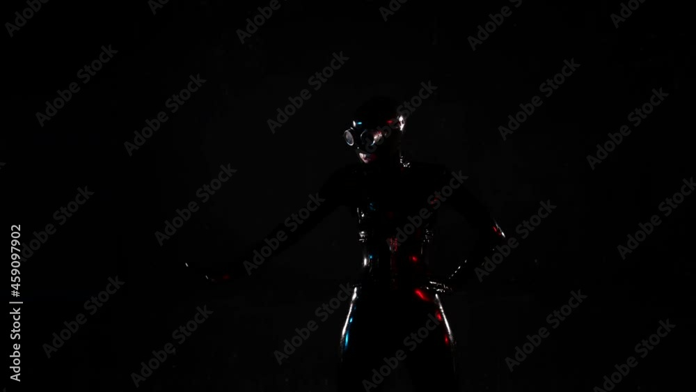 a woman in a black latex suit, helmet and glasses has one hand on belt and moves the other against a dark background. blue and red illumination. medium plan a woman in a black latex suit, helmet and glasses has one hand on belt and moves the other against a dark background. blue and red illumination. medium plan