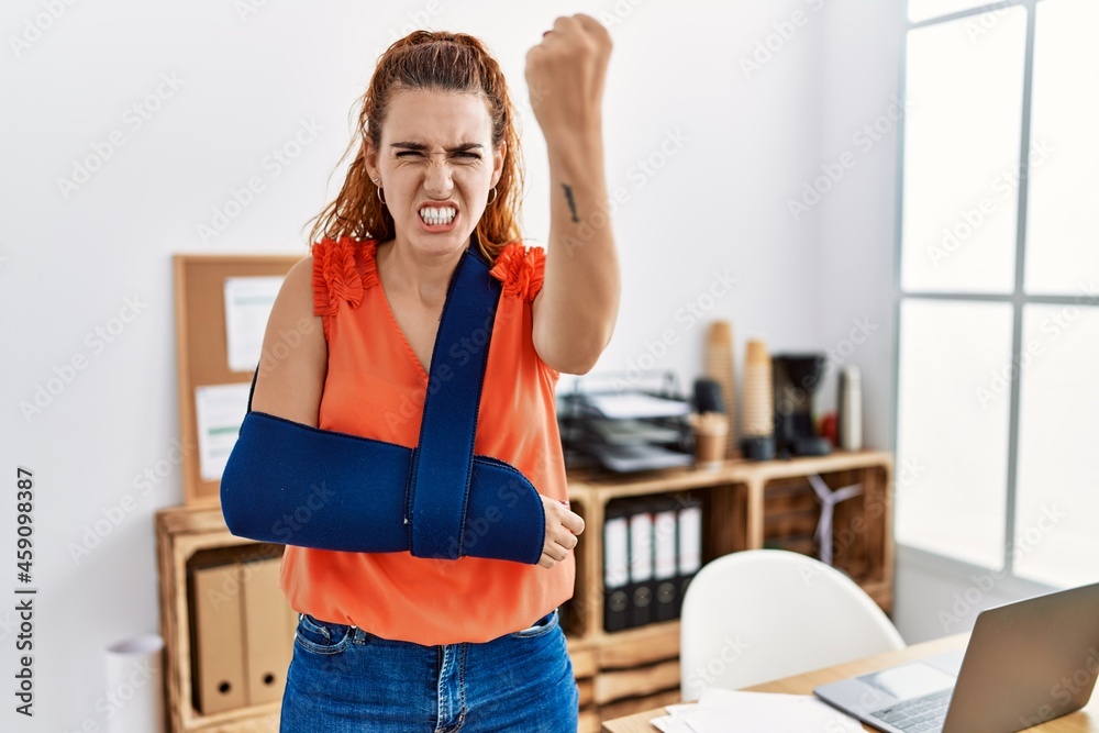 Young redhead woman wearing arm on sling at the office annoyed and frustrated shouting with anger, yelling crazy with anger and hand raised Young redhead woman wearing arm on sling at the office annoyed and frustrated shouting with anger, yelling crazy with anger and hand raised