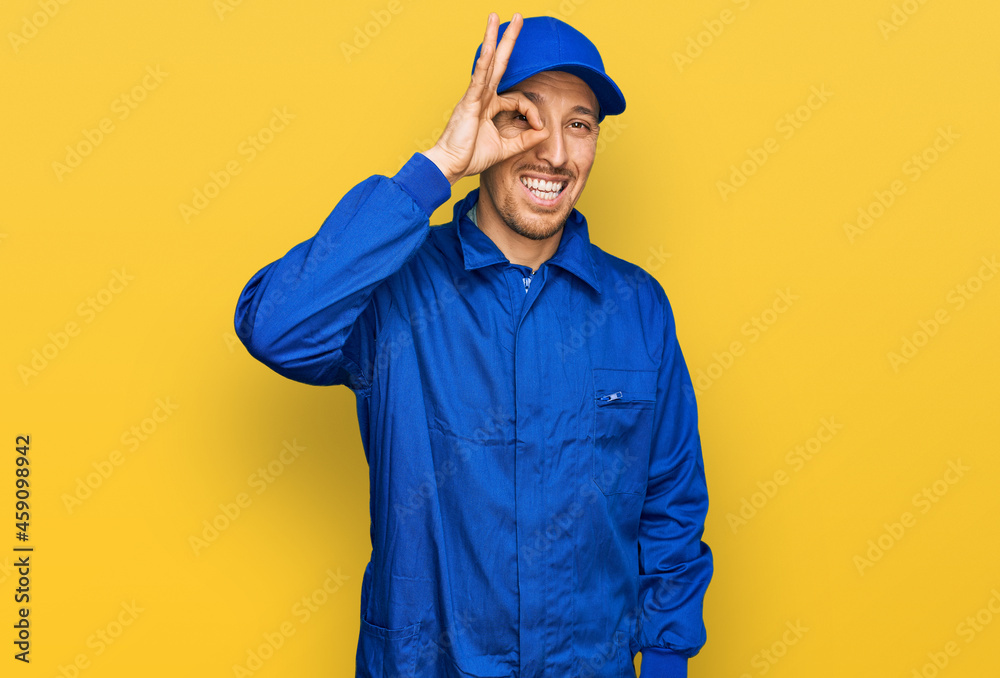 Bald man with beard wearing builder jumpsuit uniform doing ok gesture with hand smiling, eye looking through fingers with happy face.