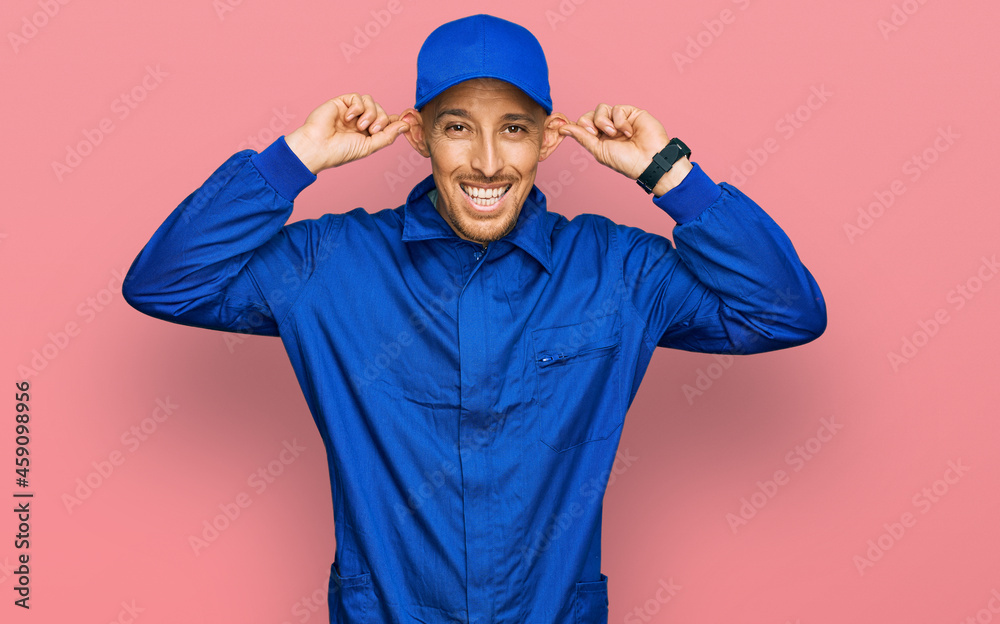 Naklejka premium Bald man with beard wearing builder jumpsuit uniform smiling pulling ears with fingers, funny gesture. audition problem