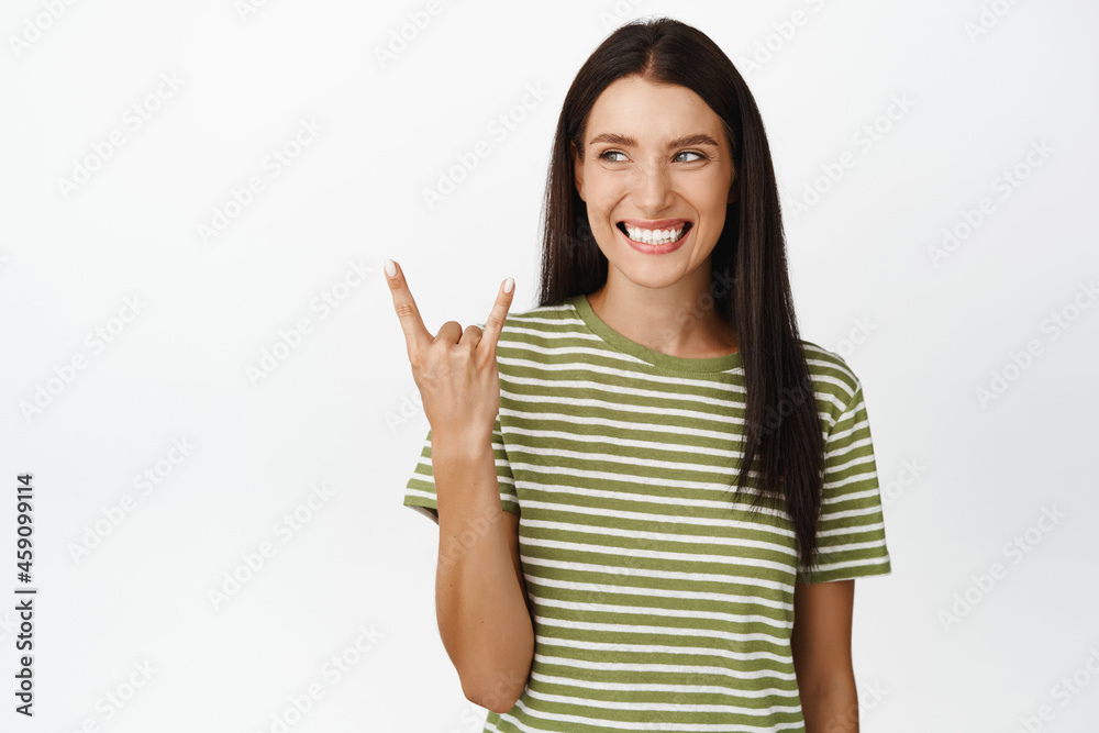 Happy brunette woman shows rock on, heavy metal gesture, smiling and looking aside at event announcement, standing over white background Happy brunette woman shows rock on, heavy metal gesture, smiling and looking aside at event announcement, standing over white background