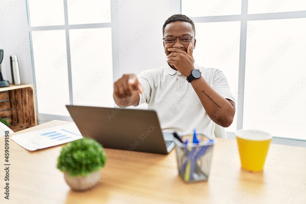 Young african man working at the office using computer laptop laughing at you, pointing finger to the camera with hand over mouth, shame expression Young african man working at the office using computer laptop laughing at you, pointing finger to the camera with hand over mouth, shame expression
