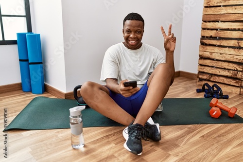 Young african man sitting on training mat at the gym using smartphone smiling with happy face winking at the camera doing victory sign with fingers. number two.