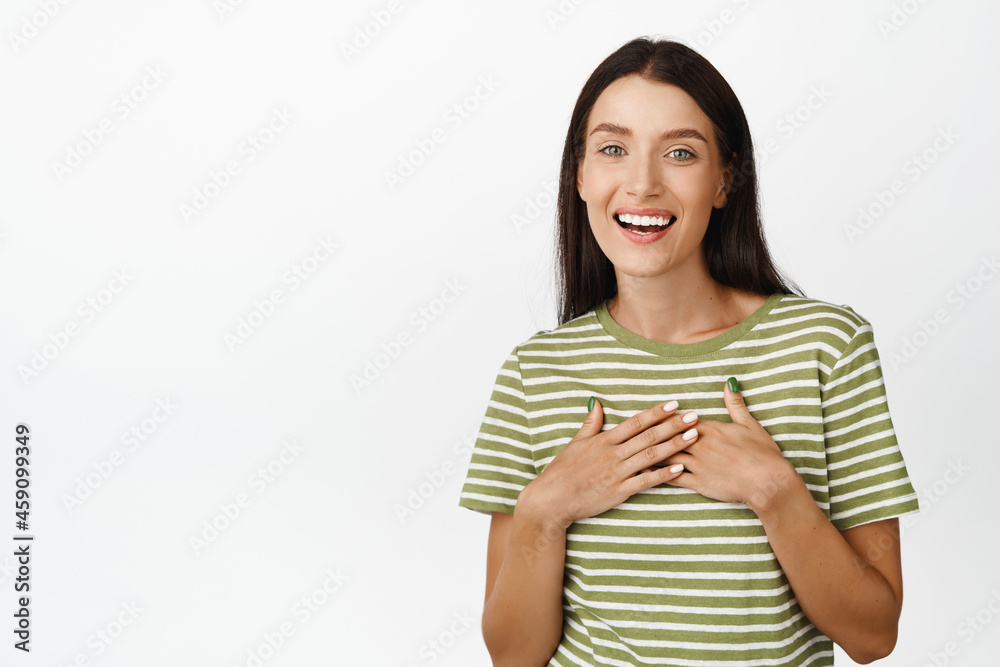 Image of pleased and thankful brunette girl look with appreciation, holding hands on chest, standing in t-shirt over white background Image of pleased and thankful brunette girl look with appreciation, holding hands on chest, standing in t-shirt over white background