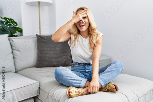 Beautiful blonde woman sitting on the sofa at home peeking in shock covering face and eyes with hand, looking through fingers with embarrassed expression.