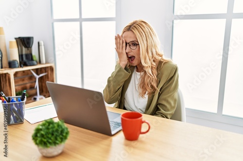 Beautiful blonde woman working at the office with laptop covering one eye with hand, confident smile on face and surprise emotion.