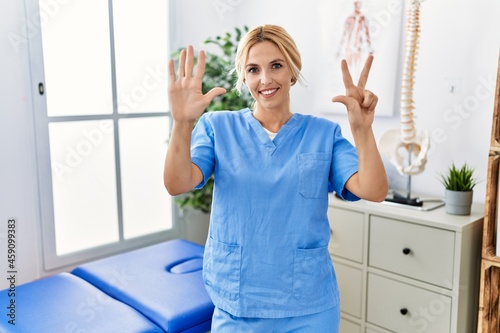 Beautiful blonde physiotherapist woman working at pain recovery clinic showing and pointing up with fingers number eight while smiling confident and happy.