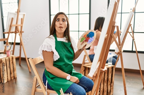 Young hispanic artist women painting on canvas at art studio making fish face with lips, crazy and comical gesture. funny expression.