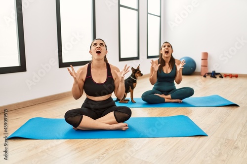 Two hispanic women friends training at the gym sitting on yoga mat with dog crazy and mad shouting and yelling with aggressive expression and arms raised. frustration concept.